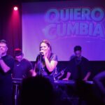 Quiero Cumbia en vivo con Euge Luján y Georgina Otero al frente, acompañados por la banda completa bajo luces violetas y el logo proyectado en pantalla.