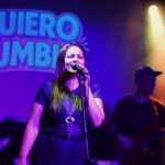 Georgina Otero cantando en vivo con Quiero Cumbia, iluminada por luces violetas y amarillas, con el logo de la banda proyectado detrás.