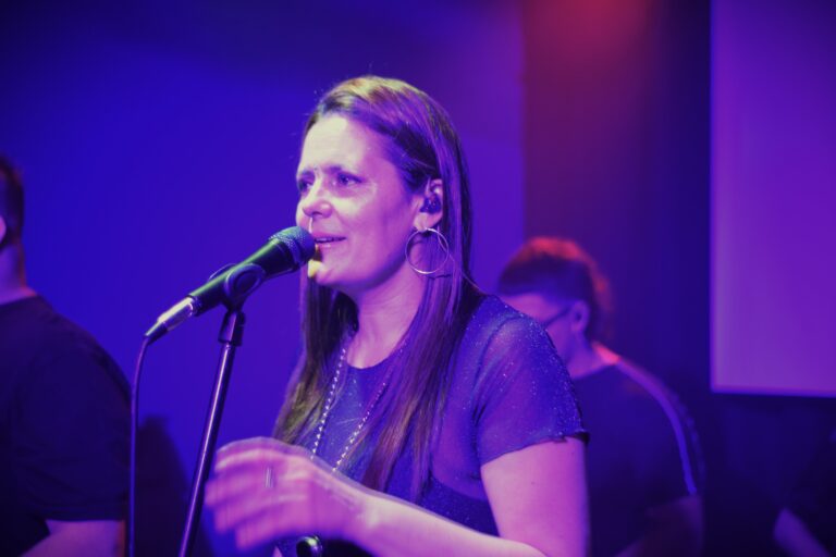 Georgina Otero cantando en vivo con Quiero Cumbia bajo luces violetas durante un show.