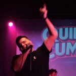 Euge Luján cantando en vivo con gesto hacia arriba y el logo de Quiero Cumbia iluminado de fondo