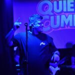 Euge Luján cantando en vivo con el logo de Quiero Cumbia iluminado de fondo