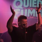 Euge Luján animando al público durante un show de Quiero Cumbia, con el logo de la banda iluminado de fondo