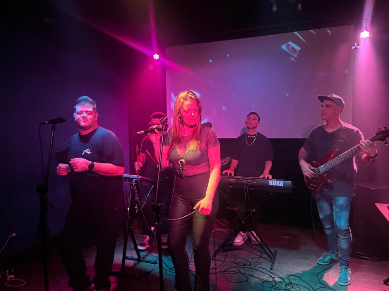 Eugenio y Georgina cantando en vivo con Quiero Cumbia junto a los músicos en teclado, percusión y bajo, bajo luces rosadas y violeta.